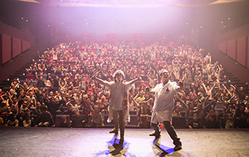 2 Mad scientists in labcoats on stage with their back towards a large excited crowd